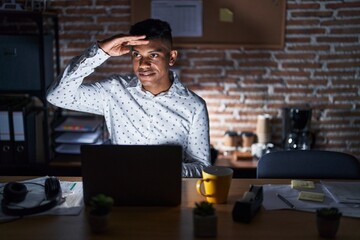 Wall Mural - Young hispanic man working at the office at night very happy and smiling looking far away with hand over head. searching concept.