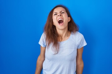 Poster - Brunette woman standing over blue background angry and mad screaming frustrated and furious, shouting with anger. rage and aggressive concept.