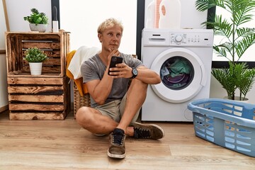 Wall Mural - Young blond man doing laundry using smartphone looking confident at the camera smiling with crossed arms and hand raised on chin. thinking positive.