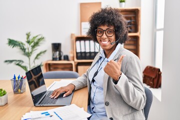 Sticker - Black woman with curly hair wearing call center agent headset at the office doing happy thumbs up gesture with hand. approving expression looking at the camera showing success.