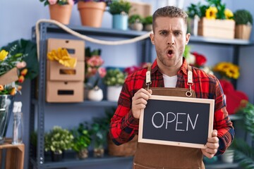 Sticker - Young caucasian man working at florist holding open sign in shock face, looking skeptical and sarcastic, surprised with open mouth