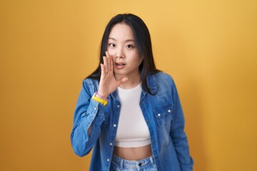 Canvas Print - Young asian woman standing over yellow background hand on mouth telling secret rumor, whispering malicious talk conversation