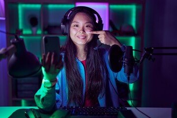 Poster - Young asian woman playing video games with smartphone pointing with hand finger to face and nose, smiling cheerful. beauty concept