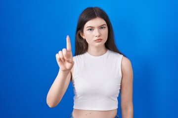 Canvas Print - Young caucasian woman standing over blue background pointing with finger up and angry expression, showing no gesture