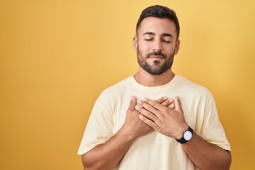 Sticker - Handsome hispanic man standing over yellow background smiling with hands on chest with closed eyes and grateful gesture on face. health concept.