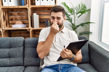 Poster - Handsome hispanic man holding clipboard working at psychology clinic smelling something stinky and disgusting, intolerable smell, holding breath with fingers on nose. bad smell