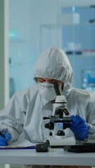 Wall Mural - Vertical video: Biologist in ppe suit working on microscope and writing information on clipboard in equipped laboratory. Chemist examining vaccine evolution using high tech researching diagnosis