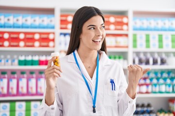 Poster - Hispanic woman working at pharmacy drugstore holding pills pointing thumb up to the side smiling happy with open mouth