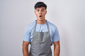 Poster - Hispanic young man wearing apron over white background afraid and shocked with surprise and amazed expression, fear and excited face.