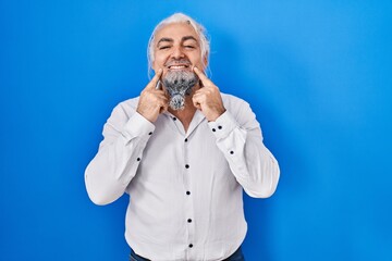 Canvas Print - Middle age man with grey hair standing over blue background smiling with open mouth, fingers pointing and forcing cheerful smile