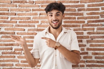 Poster - Arab man with beard standing over bricks wall background amazed and smiling to the camera while presenting with hand and pointing with finger.