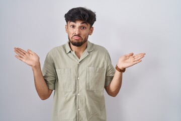 Sticker - Arab man with beard standing over white background clueless and confused expression with arms and hands raised. doubt concept.