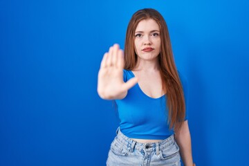 Wall Mural - Redhead woman standing over blue background doing stop sing with palm of the hand. warning expression with negative and serious gesture on the face.