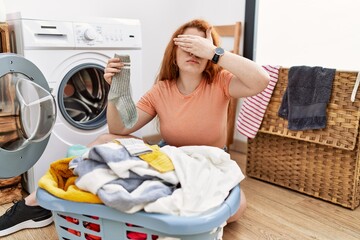 Wall Mural - Young redhead woman putting dirty laundry into washing machine covering eyes with hand, looking serious and sad. sightless, hiding and rejection concept