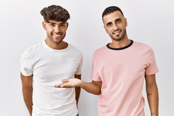 Wall Mural - Young gay couple standing over isolated background smiling cheerful presenting and pointing with palm of hand looking at the camera.