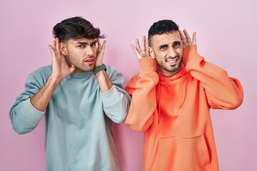 Poster - Young hispanic gay couple standing over pink background trying to hear both hands on ear gesture, curious for gossip. hearing problem, deaf