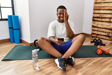 Sticker - Young african man sitting on training mat at the gym using smartphone covering one eye with hand, confident smile on face and surprise emotion.