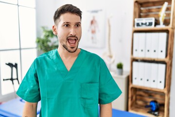 Wall Mural - Young physiotherapist man working at pain recovery clinic angry and mad screaming frustrated and furious, shouting with anger. rage and aggressive concept.