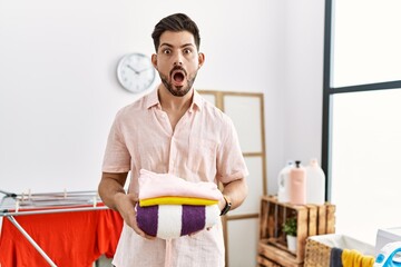 Poster - Young man with beard holding folded laundry after ironing afraid and shocked with surprise and amazed expression, fear and excited face.