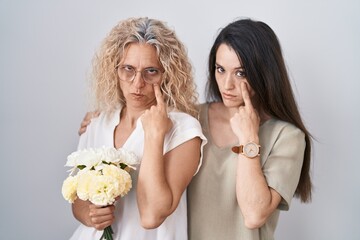 Sticker - Mother and daughter holding bouquet of white flowers pointing to the eye watching you gesture, suspicious expression