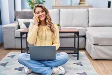 Wall Mural - Young woman using laptop and talking on smartphone at home