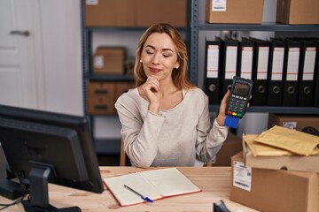 Wall Mural - Hispanic woman working at small business ecommerce holding credit card and dataphone serious face thinking about question with hand on chin, thoughtful about confusing idea