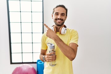 Sticker - Young hispanic man wearing sportswear and drinking water at the gym cheerful with a smile on face pointing with hand and finger up to the side with happy and natural expression