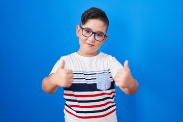 Sticker - Young hispanic kid standing over blue background success sign doing positive gesture with hand, thumbs up smiling and happy. cheerful expression and winner gesture.