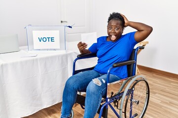 Poster - Young african woman sitting on wheelchair voting putting envelop in ballot box crazy and scared with hands on head, afraid and surprised of shock with open mouth