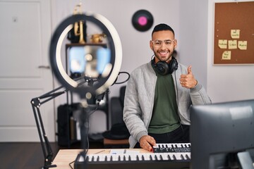 Wall Mural - Young hispanic man playing piano at music studio recording himself smiling happy and positive, thumb up doing excellent and approval sign