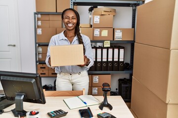 Canvas Print - African woman working at small business ecommerce holding box winking looking at the camera with sexy expression, cheerful and happy face.