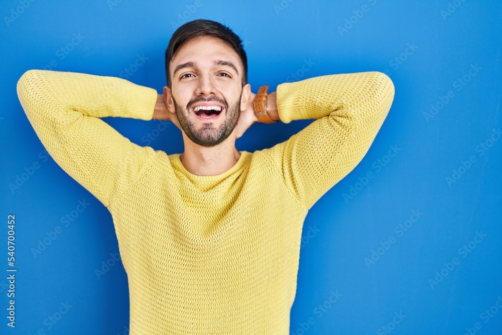 Hispanic man standing over blue background relaxing and stretching, arms and hands behind head and neck smiling happy