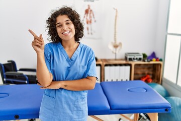 Canvas Print - Young hispanic physiotherapist woman working at pain recovery clinic smiling happy pointing with hand and finger to the side