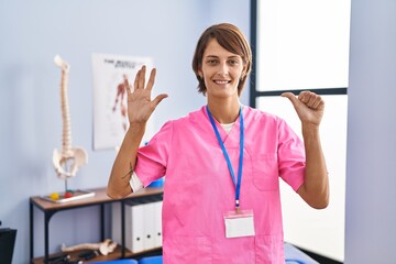 Poster - Brunette woman working at rehabilitation clinic showing and pointing up with fingers number six while smiling confident and happy.