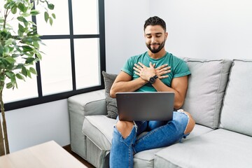 Poster - Young handsome man with beard using computer laptop sitting on the sofa at home smiling with hands on chest with closed eyes and grateful gesture on face. health concept.