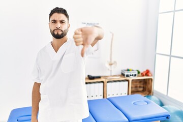 Canvas Print - Young handsome man with beard working at pain recovery clinic looking unhappy and angry showing rejection and negative with thumbs down gesture. bad expression.