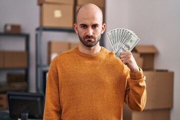 Canvas Print - Young bald man with beard working at small business ecommerce holding money thinking attitude and sober expression looking self confident