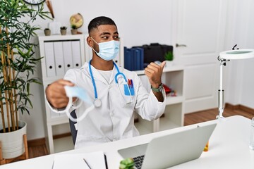 Wall Mural - Young indian doctor offering safety mask pointing thumb up to the side smiling happy with open mouth