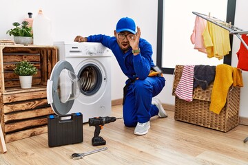 Wall Mural - Young indian technician working on washing machine doing ok gesture shocked with surprised face, eye looking through fingers. unbelieving expression.