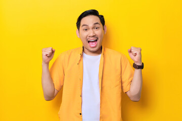 Excited young handsome Asian man celebrating success with raised fists isolated on yellow background