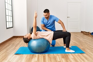 Wall Mural - Latin man and woman wearing physiotherapist uniform having rehab session using fit ball at rehab center