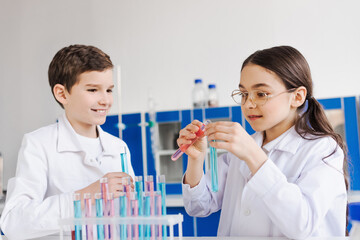 Canvas Print - girl in eyeglasses mixing liquid in test tubes near smiling friend in white coat.