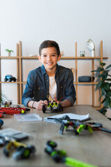 Canvas Print - cheerful boy looking at camera near mechanical details of robotics model on table at home.