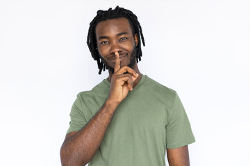 Wall Mural - Portrait of pleased African American man shushing. Secretive young male model with braided dark hair in green T-shirt looking at camera with finger on lips, asking to be quiet. Secret, silence concept