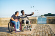 Happy biracial couple taking selfie on embankment. African American man and Caucasian woman in wheelchairs on embankment, holding mobile phone, smiling. Love, relationship, social media concept