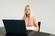© dikushin - Studio portrait of charming blonde young woman with broken arm wrapped in plaster bandage sitting at black table with laptop computer, looking at camera, on white isolated background.