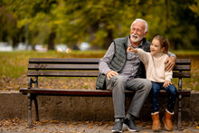 Grandfather Spending Time With His Granddaughter On Bench In Park On Autumn Day