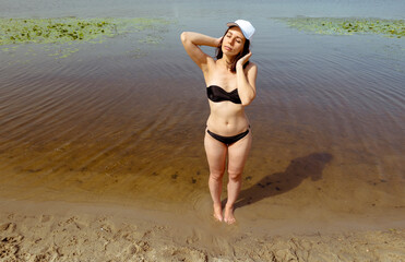 A woman in a black swimsuit and a white cap stands in the water near the beach. Young woman closes her eyes while relaxing in summer. Woman portrait beach