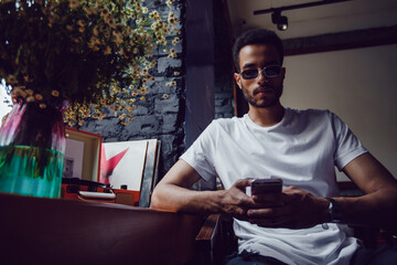 Wall Mural - African american man in a white t-shirt sits in a coffee shop and uses the phone. Mock-up.