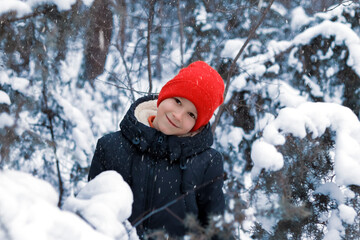 Wall Mural - A happy smiling boy in a black down jacket and a red knitted hat plays and runs in a beautiful snowy winter park for Christmas